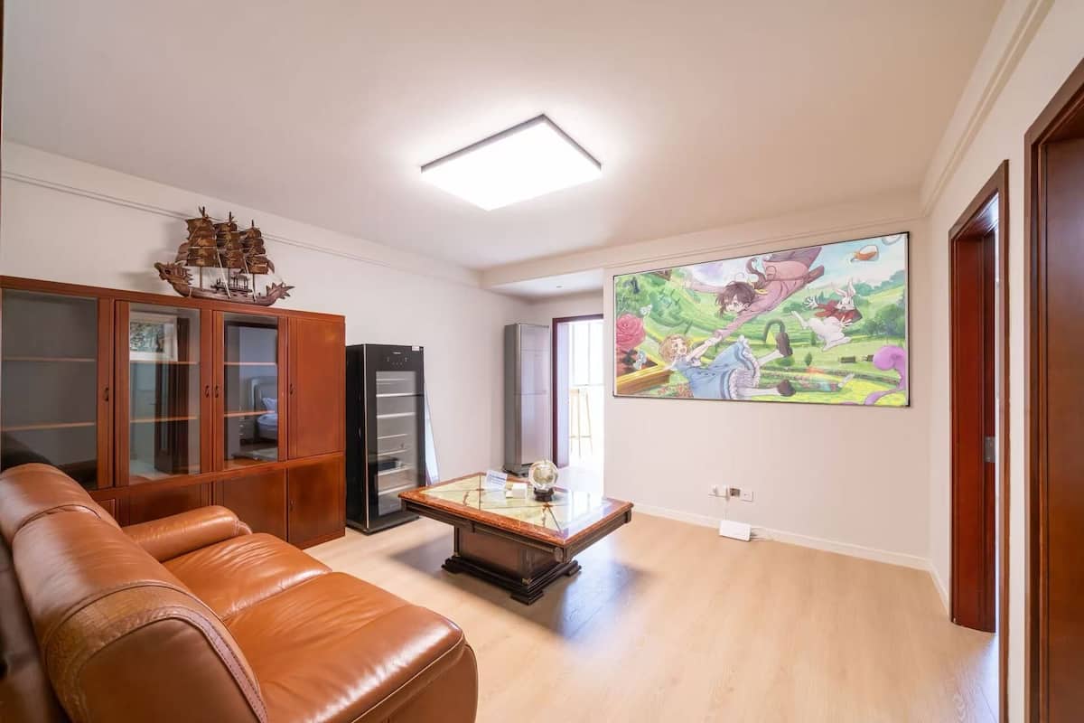 Una sala de estar con un sofá de cuero marrón, un mueble de madera, un refrigerador negro, una mesa de centro de vidrio y un cuadro grande de estilo anime en la pared.