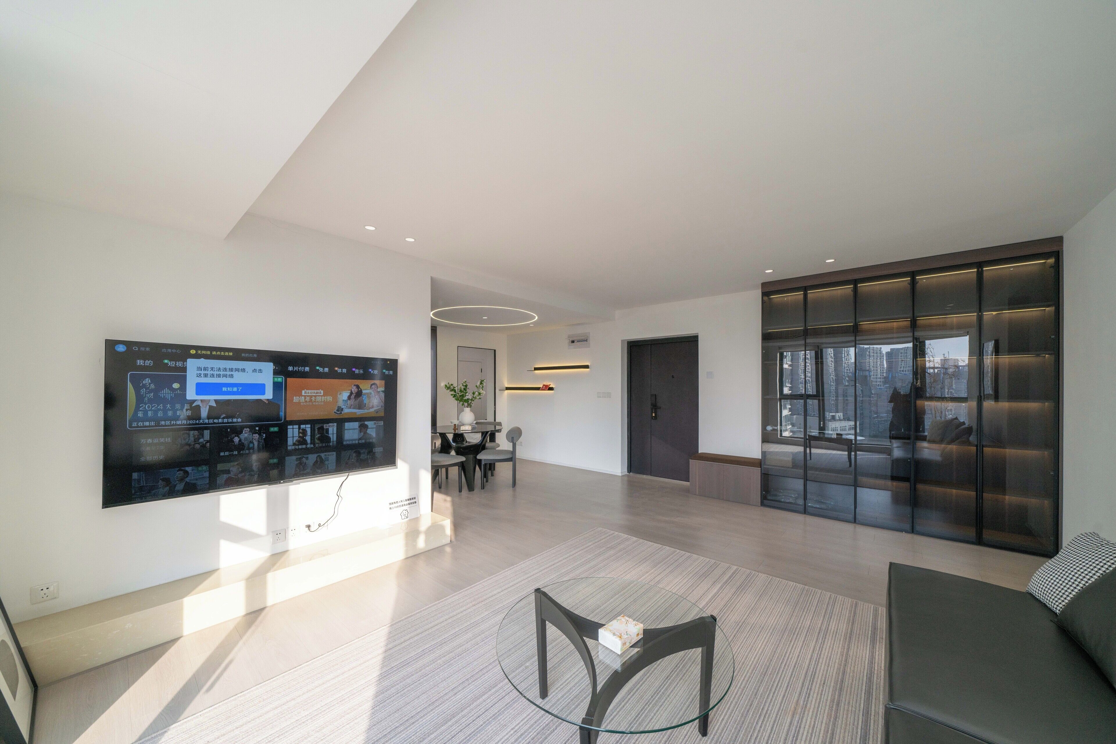A modern living space with a flat-screen TV mounted on the wall, a glass coffee table, a sofa, and a large window offering a view of the cityscape.
