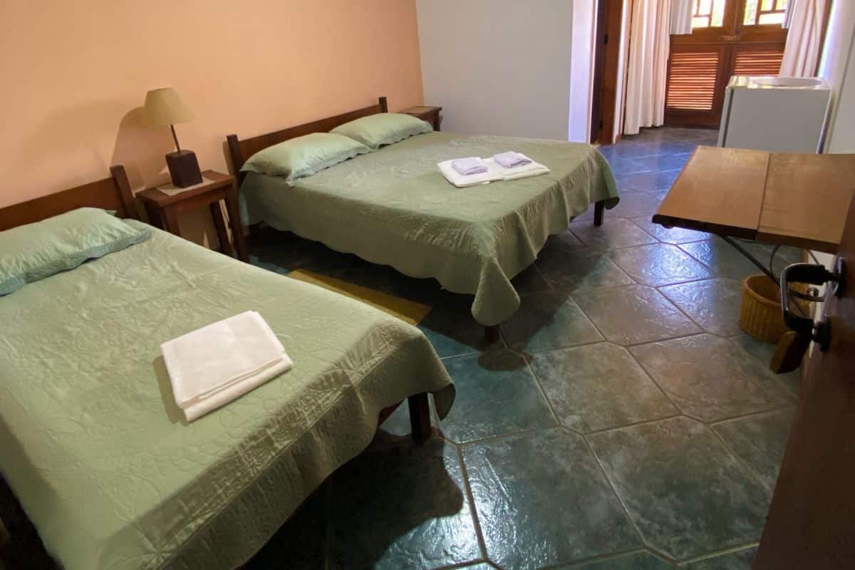 Quarto com duas camas, um ventilador de teto, uma luminária e uma janela com cortinas.