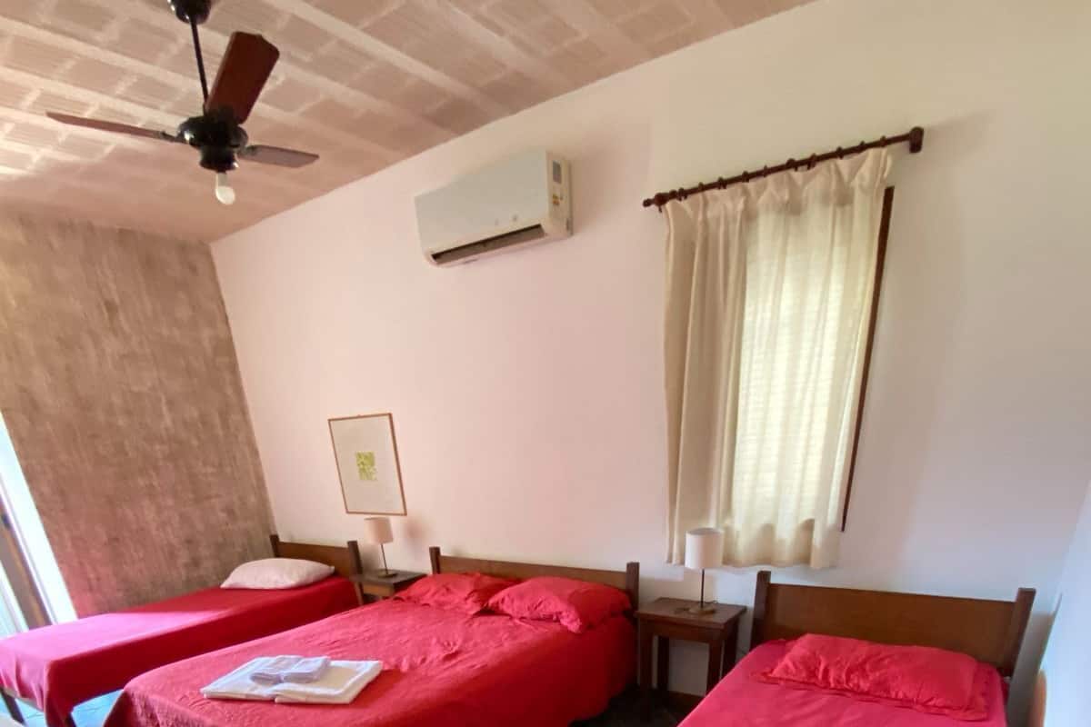 Quarto com três camas, cada uma com colcha vermelha, ventilador de teto, ar condicionado e janela com cortinas.