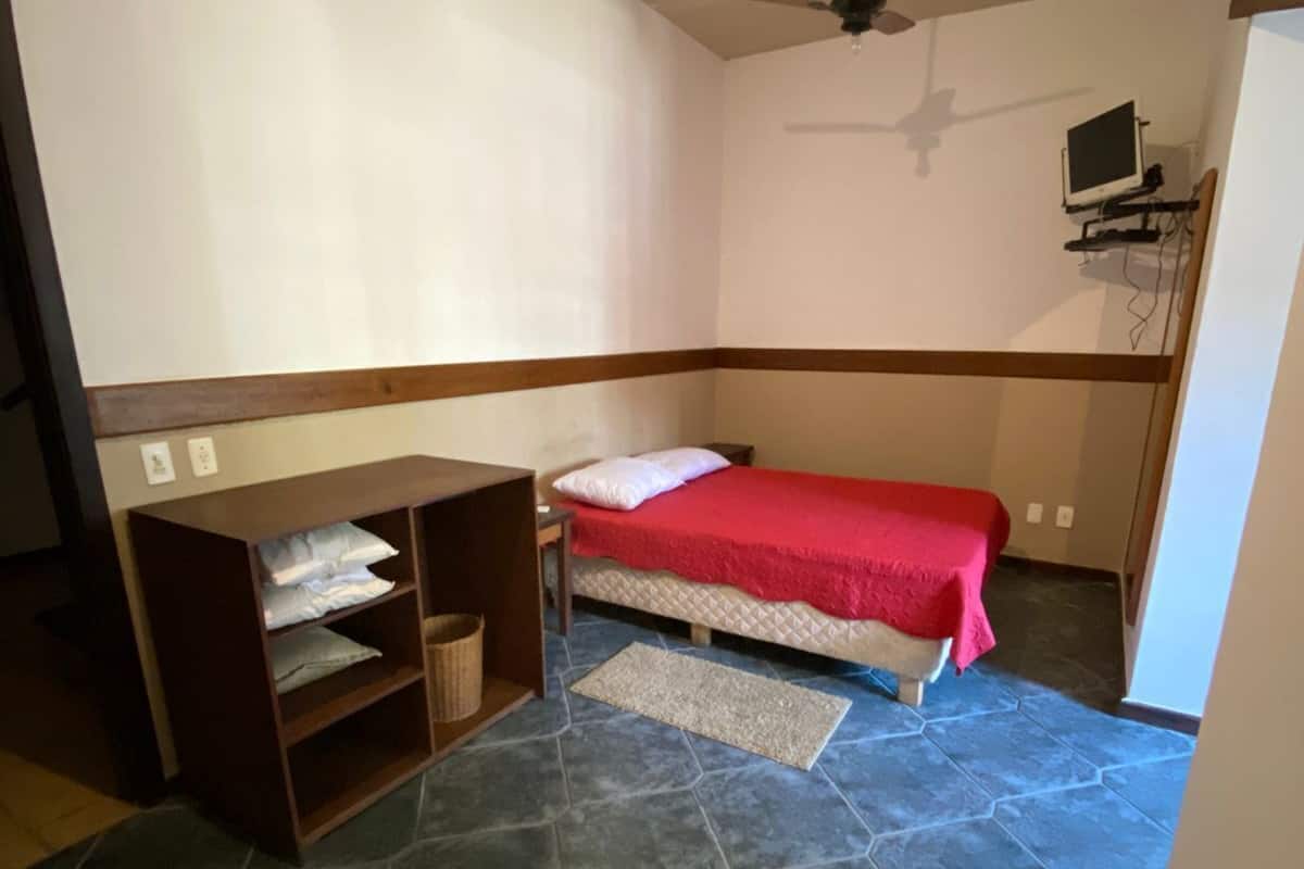 Quarto com cama, escrivaninha, cadeira, ventilador de teto e televisão sobre suporte.