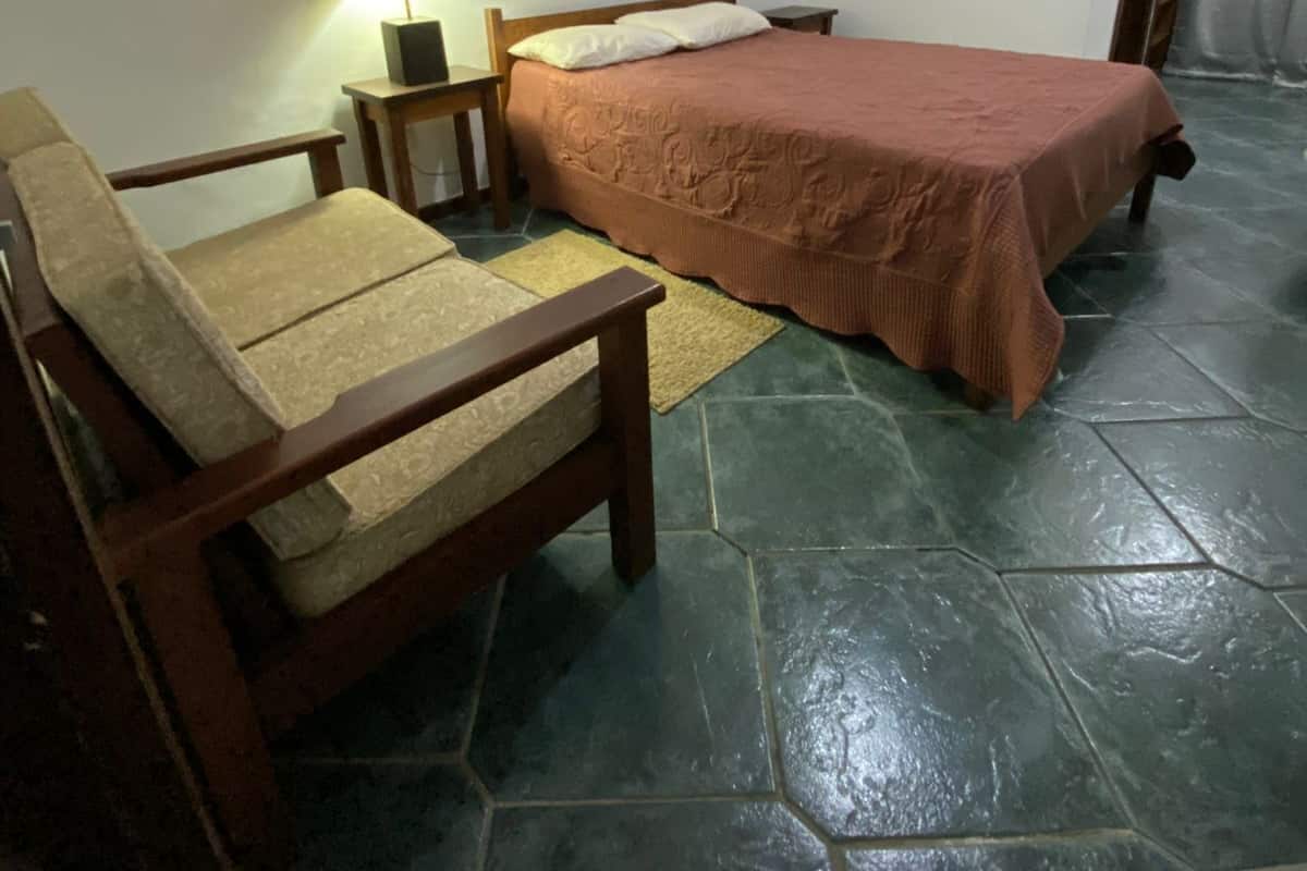 Quarto com uma cama, uma cadeira de madeira, mesa de cabeceira com luminária e piso ladrilhado.