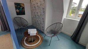 A room with a blue carpet, a round wooden table with a white towel, two wire-framed chairs, a patterned wall, and a window with curtains. - La Bonnerie - The Dream Observatory (La Bouëxière)