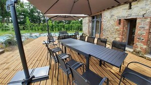 Un patio al aire libre con una mesa larga y sillas, un paraguas grande y un edificio de piedra al fondo.