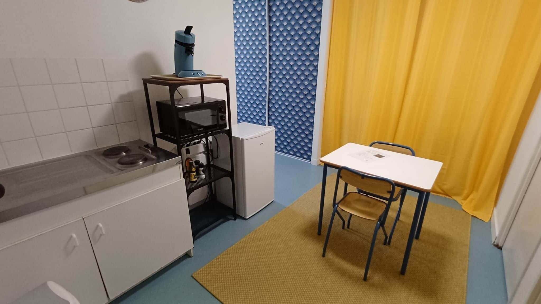 Een kleine keuken met een kookplaat, koelkast en een tafeltje met stoelen.