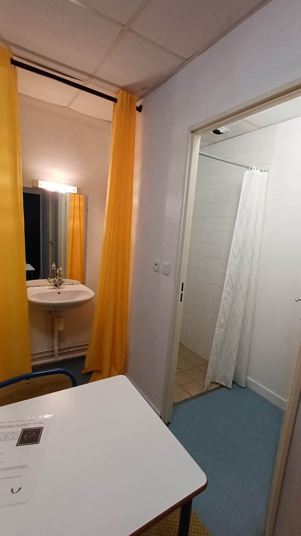 Een kleine kamer met een witte tafel, een wastafel met spiegel en een douchecabine met een witte gordijn.