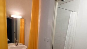 Una habitación pequeña con una mesa blanca, un lavabo con espejo y una zona de ducha con cortina blanca.