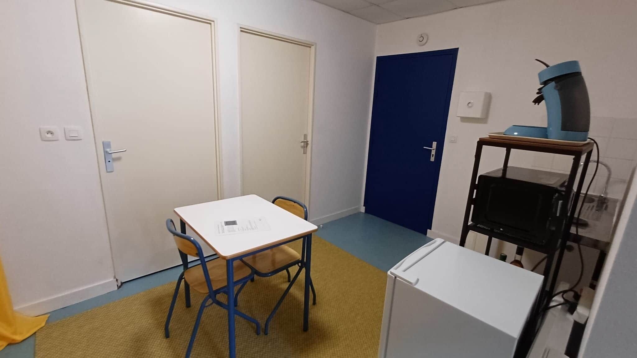 Een kleine kamer met een witte tafel, twee stoelen, een blauwe deur en een magnetron.