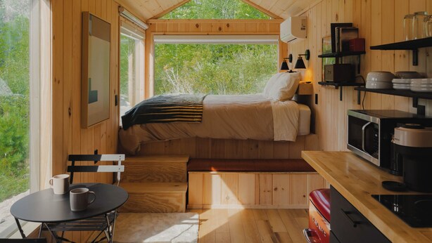 A wooden cabin interior with a bed, kitchen, dining area, and large windows.