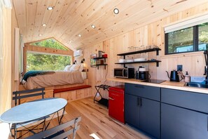 Een compact vakantiehuisje met een bed, kitchenette, eethoek en een raam met uitzicht op groen.