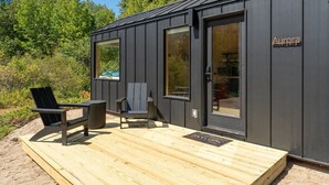A modern cabin with a wooden deck, two Adirondack chairs, and a glass door labeled 'Aurora.'