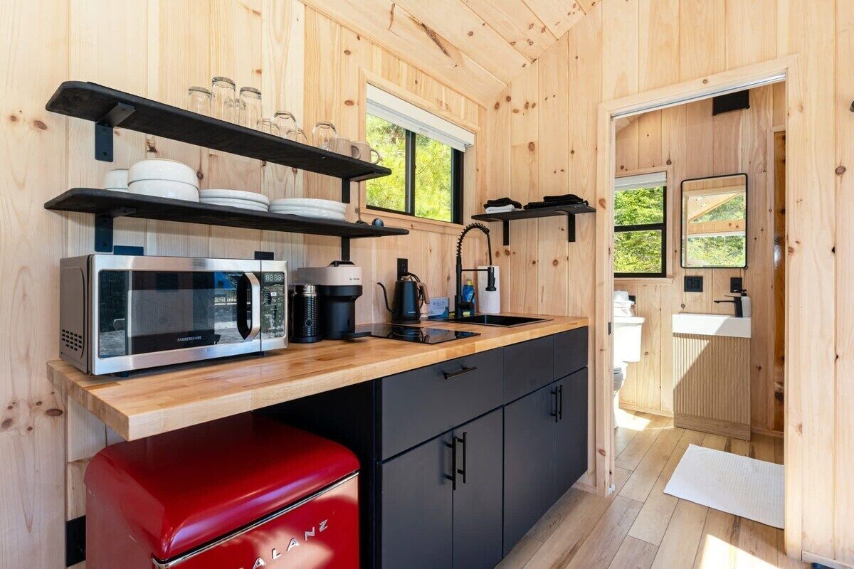 A compact kitchen with a microwave, coffee maker, and a red appliance, wooden walls, and a window.