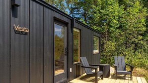 A modern wooden cabin with a 'Vesta' sign, a deck with two chairs and a table, and a door with a 'Welcome' mat. - Vesta Nook! With Shower, Queen Bed & pullout sofa (Sullivan)
