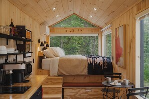 A wooden cabin with a bed, a small table with cups, a coffee machine, and a view of the forest through a large window. - Vesta Nook! With Shower, Queen Bed & pullout sofa (Sullivan)