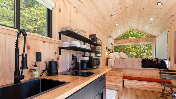 A compact kitchen with a sink, stove, and shelves, a cozy seating area with a couch and table, and a large window offering a view of trees. - Vesta Nook! With Shower, Queen Bed & pullout sofa (Sullivan)