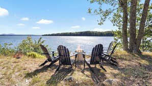 Vista para o lago com duas cadeiras Adirondack de frente para uma lareira, rodeado por árvores e céu claro.