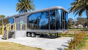 A modern, compact mobile home with a large sliding door, a small porch, and a staircase leading to the entrance.