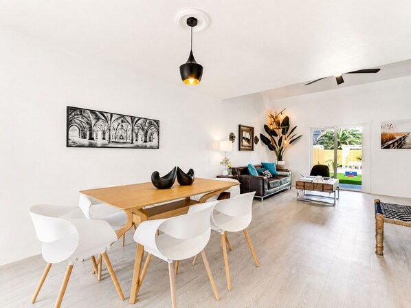 A modern living room with a dining area, featuring a wooden table, white chairs, a sofa, and a large plant.