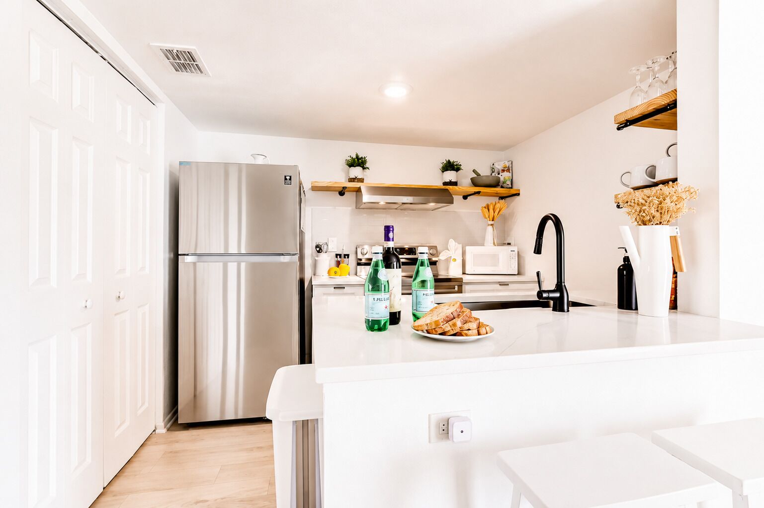 A modern kitchen with a stainless steel refrigerator, a microwave, and a sink with a black faucet. There are bottles of water and a plate of bread on the counter.