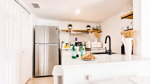 A modern kitchen with a stainless steel refrigerator, a microwave, and a sink with a black faucet. There are bottles of water and a plate of bread on the counter. - Classy Bohemian Beach Cabana- Close to Beach (St. Augustine)