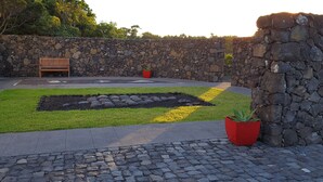 Une cour avec un mur en pierre, un banc, un jardin agrémenté de rochers et de fleurs jaunes, et un bac à fleurs rouge.