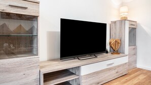 A modern living room with a flat-screen TV on a wooden TV stand, a glass cabinet, and a small table with a lamp. - Apartment 'Zum Waldblick' with Shared Garden and Wi-Fi (Wulsbüttel )