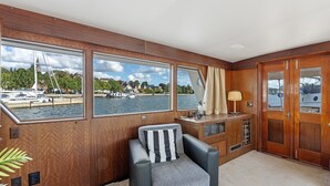 Interior de un yate con vistas al puerto deportivo, una zona de estar con cojines, una pequeña mesa con lámpara y una puerta de madera.