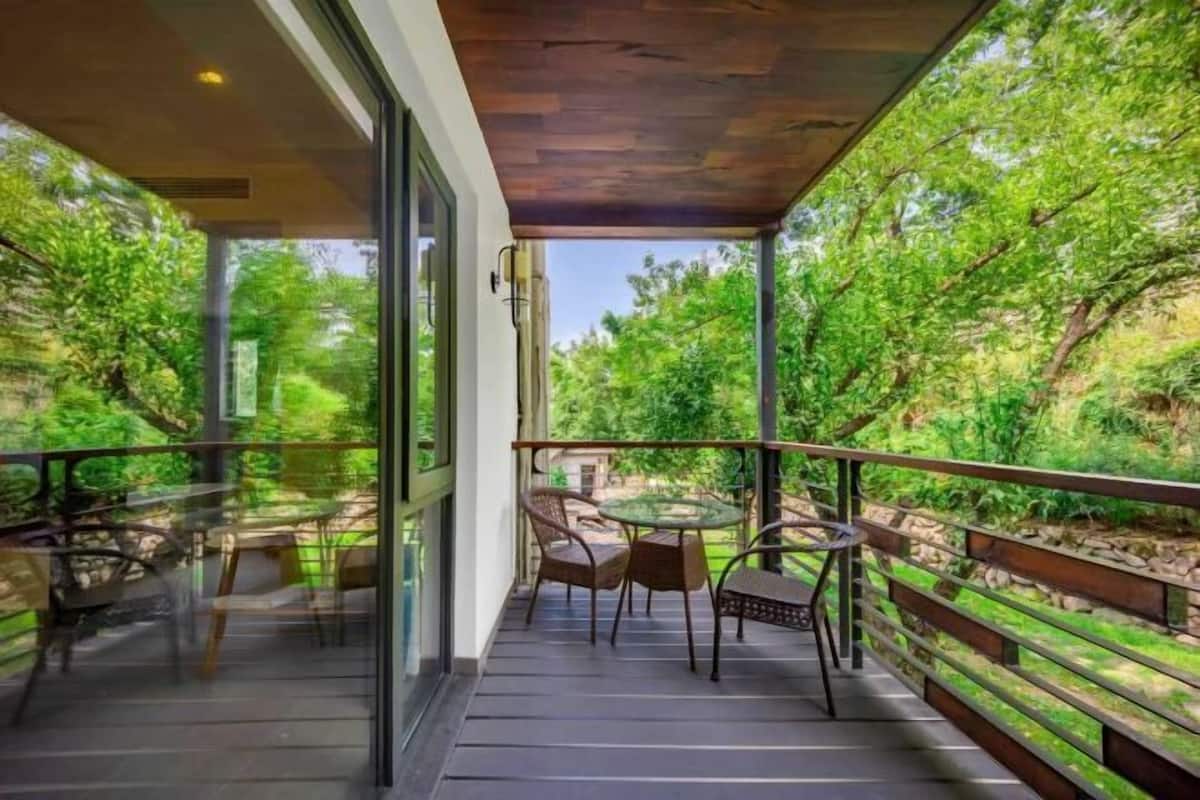A balcony with a glass door, a table, chairs, and a wooden railing overlooking a lush green landscape.