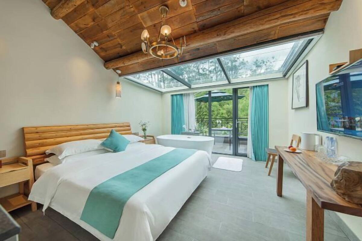 A bedroom with a wooden ceiling, a bed with white bedding and a teal throw, a round table, and a large window with curtains.