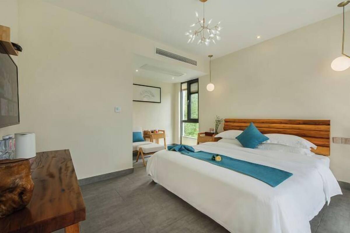 A modern hotel room with a large bed, a wooden headboard, a desk, and a view of the outdoors.