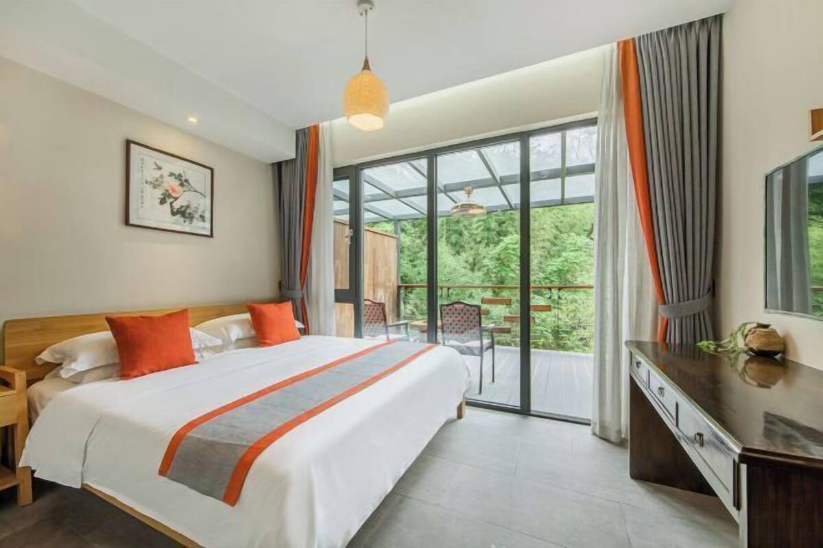 A hotel room with a large bed, a desk, a chair, and a balcony with a view of greenery.