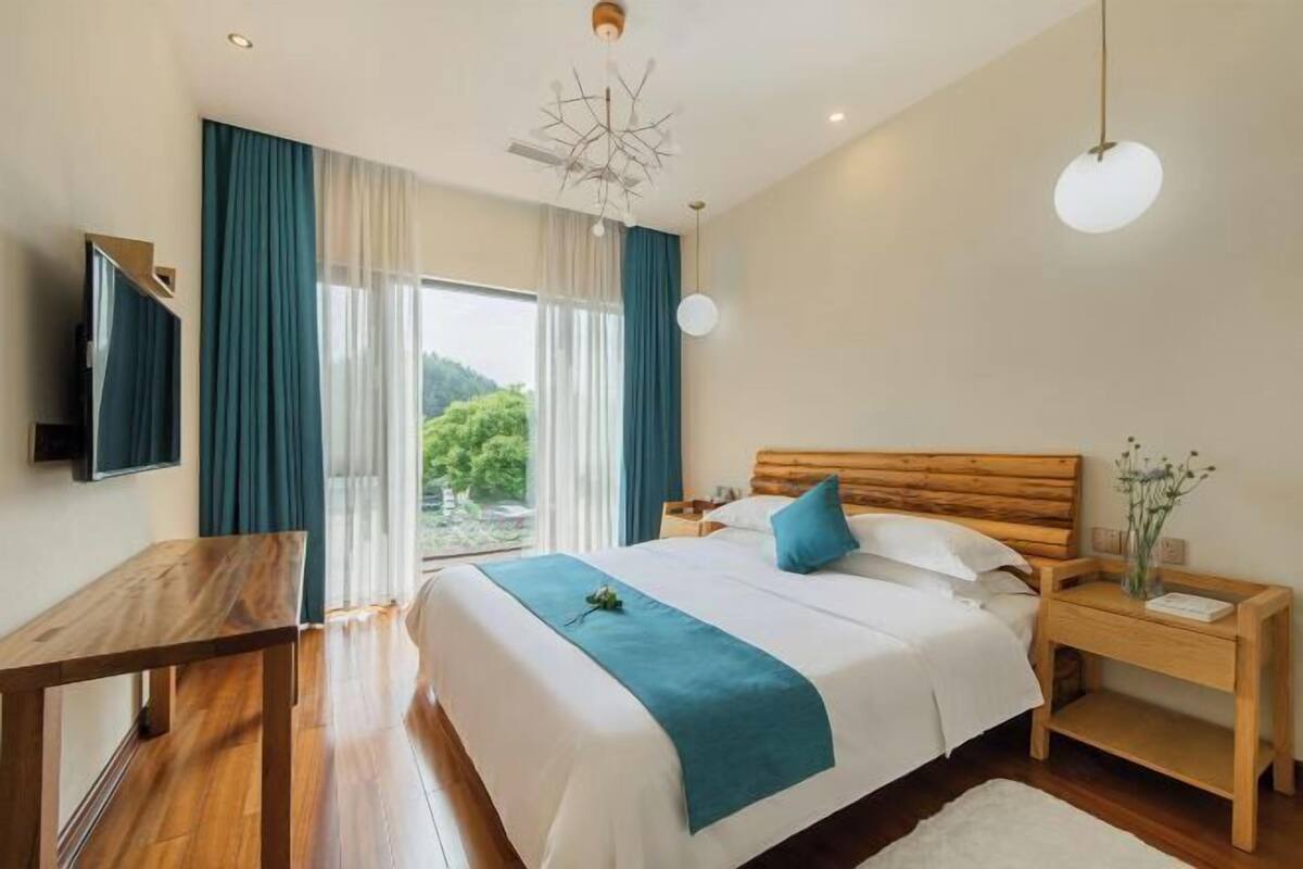 A bedroom with a wooden bed, a TV mounted on the wall, a wooden nightstand, and a large window with curtains.