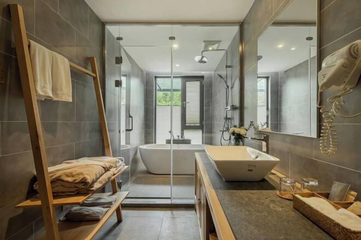 A modern bathroom with a freestanding bathtub, a large mirror, and a wooden shelf with towels.