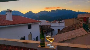 Una terraza con una botella de vino y copas, con vistas a un pueblo de edificios tradicionales y montañas al fondo.