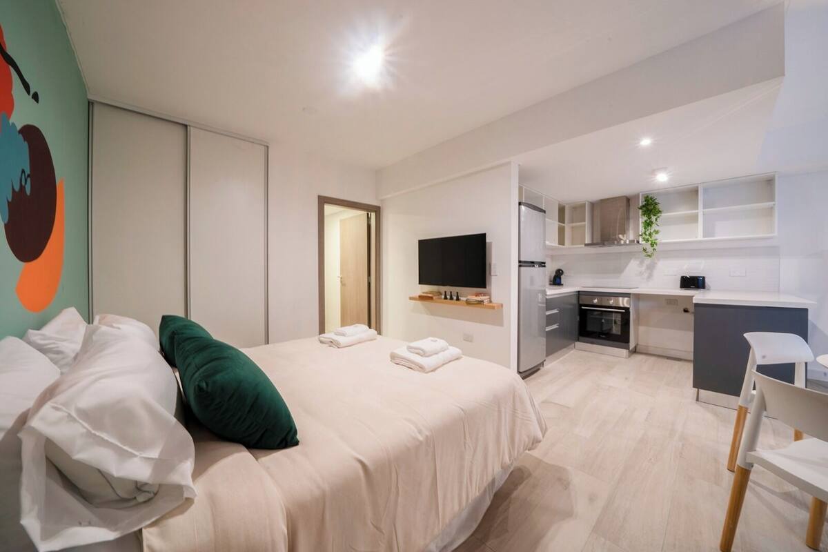 A modern hotel room with a large bed, a desk, a kitchenette, and a TV.