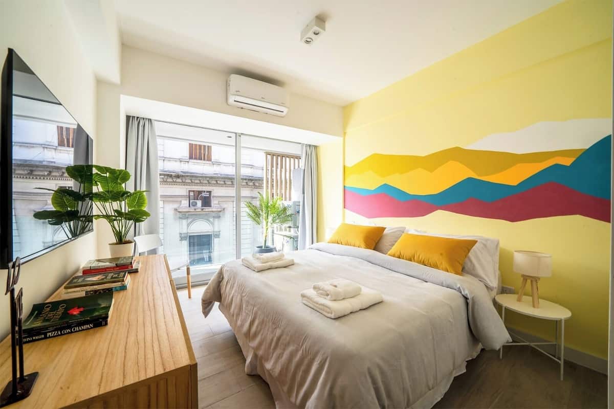 A hotel room with a bed, a desk with books, a plant, and a colorful wall mural.