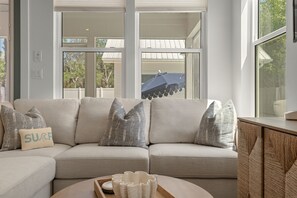 A beige sectional sofa with patterned pillows, a round wooden side table, and a view of a house through large windows. - Trae Bien – Private Pool / Pet Friendly / Golf Cart (Santa Rosa Beach)