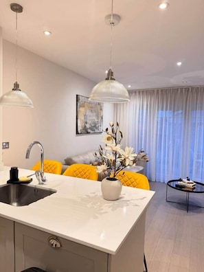 A modern kitchen with a central island, a sink, and a dining area with yellow chairs. There is a vase with flowers on the island. - Spacious 4BR Home | 3 Parking | Offers Weekly Stay (Reading)