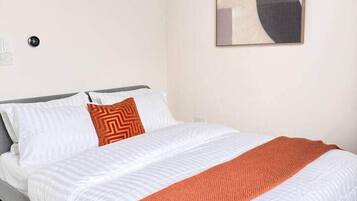 A bedroom with a bed, white bedding, an orange throw blanket, and a geometric wall art piece.