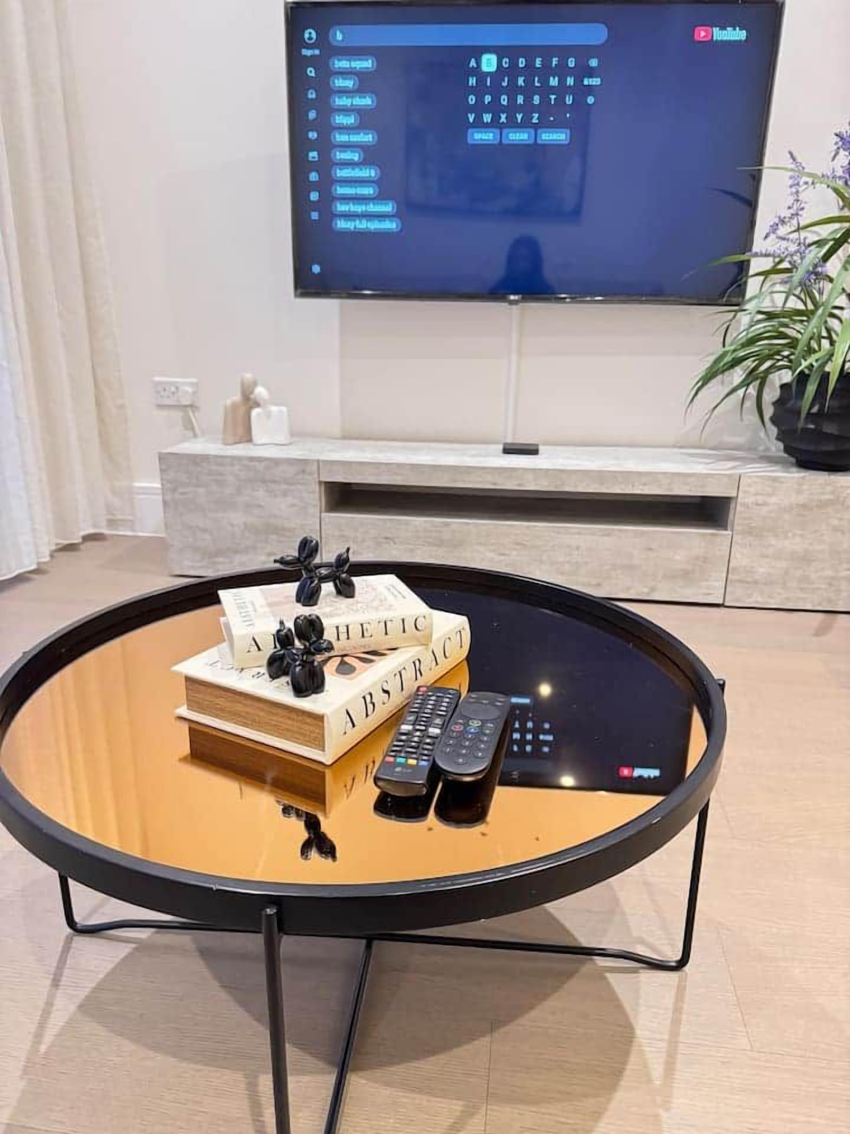 Une table basse ronde avec un plateau en verre, une pile de livres et deux télécommandes.