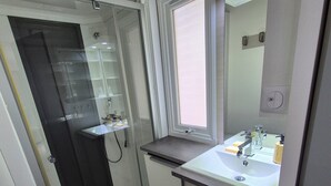 Une petite salle de bain dotée d’une cabine de douche en verre, d’un lavabo avec un miroir et d’un petit panier en osier.