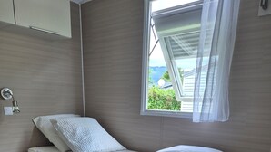 Un lit simple avec un cadre en bois, une table de chevet, une lampe fixée au mur et une vue sur la verdure par la fenêtre.