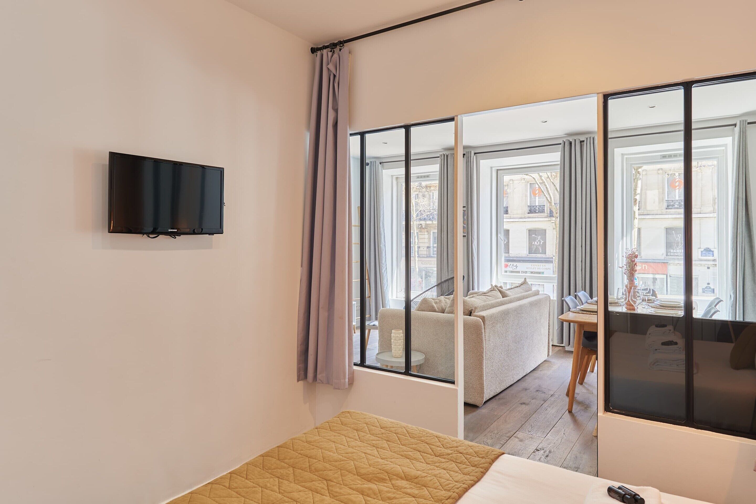 A modern hotel room with a bed, a flat-screen TV mounted on the wall, and large windows offering a view of the city.