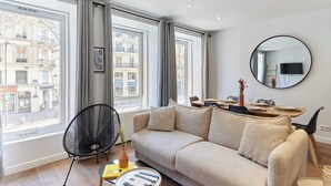 Una sala de estar moderna con un sofá, una mesa de comedor con sillas, un espejo redondo y ventanas amplias con cortinas.