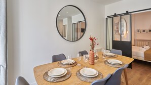Un comedor con una mesa de madera dispuesta para cuatro personas, sillas alrededor, un espejo en la pared y una cama con puerta corrediza al fondo.