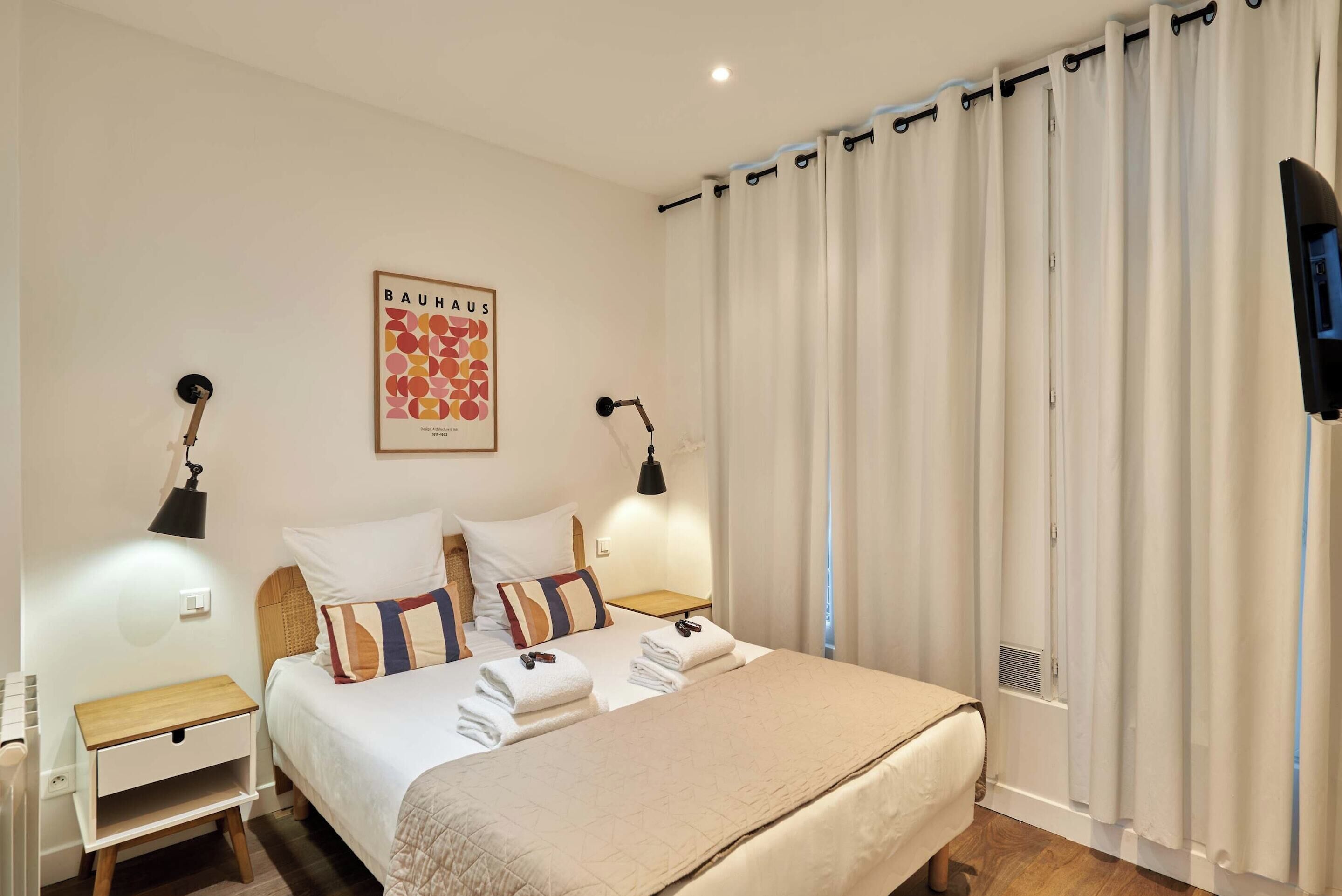 A hotel room with a bed, two bedside tables, a wall-mounted TV, and a window with curtains.