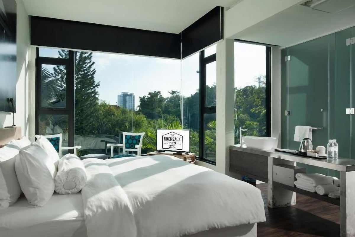 A modern hotel room with a large bed, a desk, and a view of greenery and buildings.