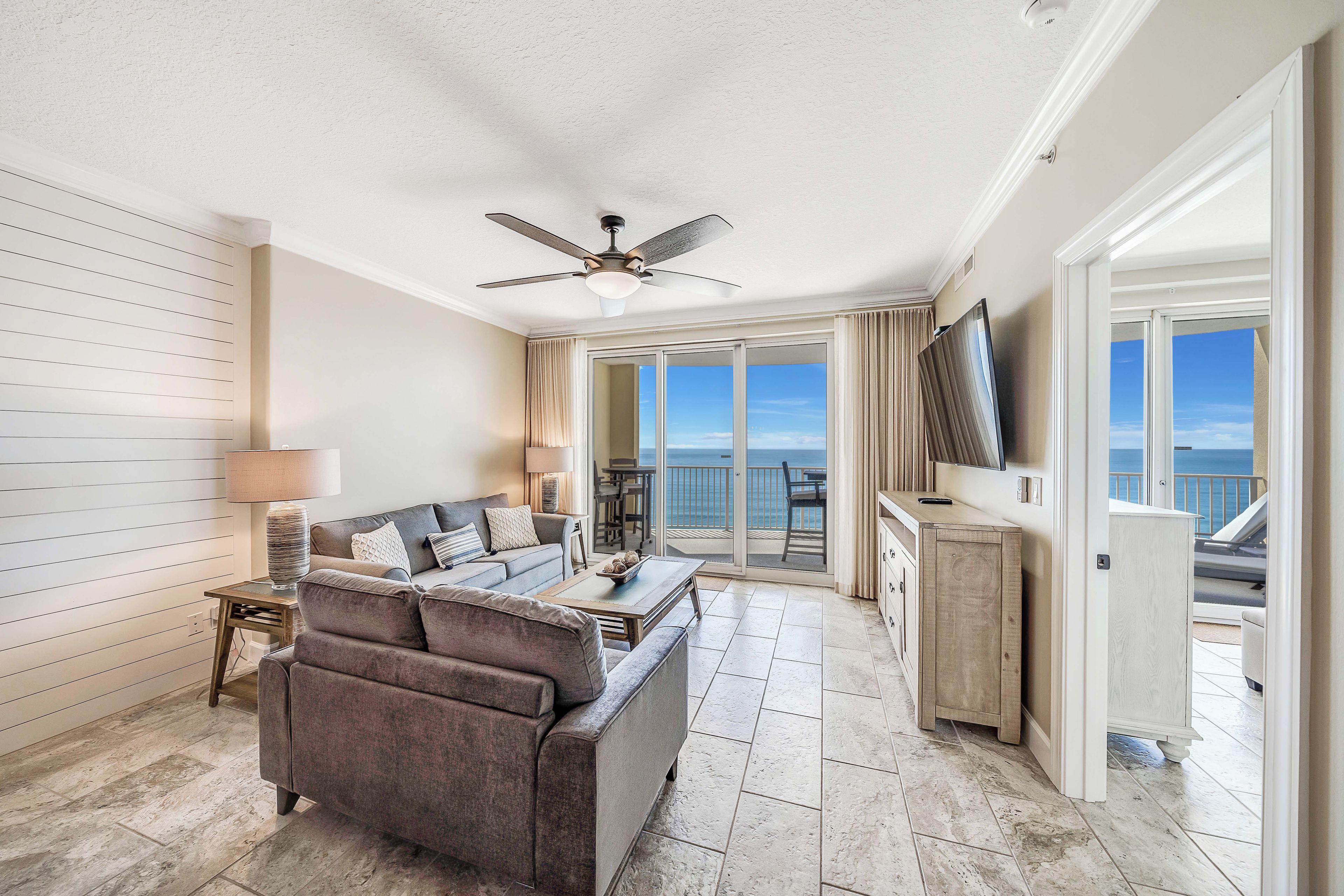A spacious living area with a sofa, coffee table, and a ceiling fan. A large window offers a view of the ocean.