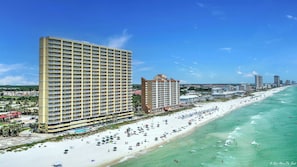 Un hotel frente a la playa con varios pisos, piscina y una playa con sombrillas y gente.