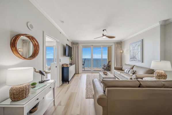 A modern living room with a sofa, a coffee table, and a TV. There is a balcony with a view of the sea.